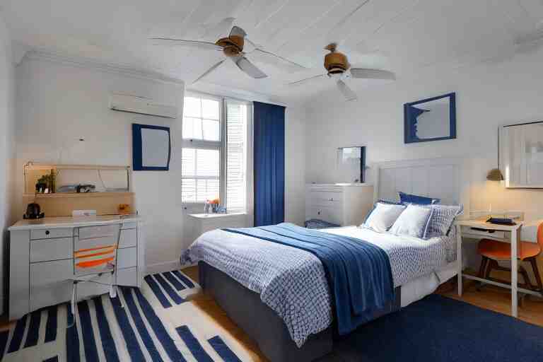 a 10 by 11 foot old bed room with blue grey criss cross patterned walls, white ceiling, navy blue carpet, a small bed, desk, two wooden wardrobes, an old TV, and a ceiling fan gives off a dim orange light at night time