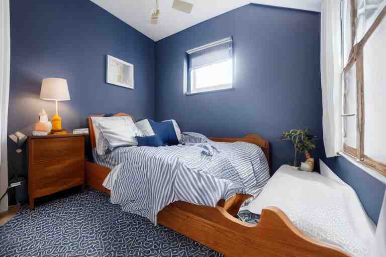 a 10 by 11 foot old bed room with blue grey criss cross patterned walls, white ceiling, navy blue carpet, a small bed, desk, two wooden wardrobes, an old TV, and a ceiling fan gives off a dim orange light at night time