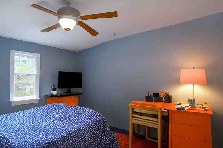 a 10 by 11 foot old bed room with blue grey criss cross patterned walls, white ceiling, navy blue carpet, a small bed, desk, two wooden wardrobes, an old TV, and a ceiling fan gives off a dim orange light at night time